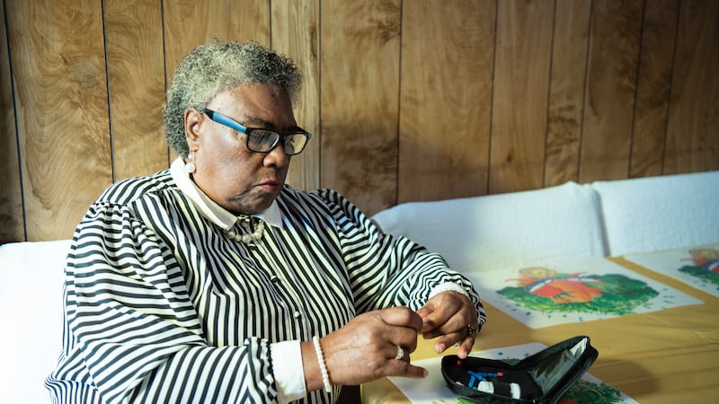 A Black woman sits at a kitchen table with a glucose monitoring kit on the table in front of...